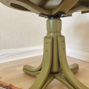 May include: A vintage, round table with a pedestal base. The table is painted in a muted olive green color. The base has four curved legs that extend outwards. The underside of the tabletop is visible, showing the wooden construction and hardware.