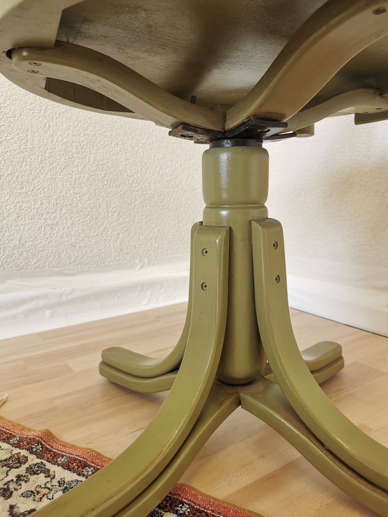 May include: A vintage, round table with a pedestal base. The table is painted in a muted olive green color. The base has four curved legs that extend outwards. The underside of the tabletop is visible, showing the wooden construction and hardware.