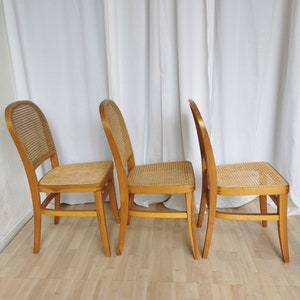 May include: Three wooden chairs with woven cane seats and backs. The chairs are arranged in a row, with the backs facing the viewer. The chairs are light brown in color and have a simple, classic design.