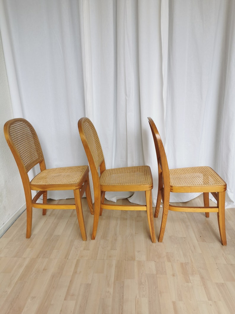 May include: Three wooden chairs with woven cane seats and backs. The chairs are arranged in a row, with the backs facing the viewer. The chairs are light brown in color and have a simple, classic design.