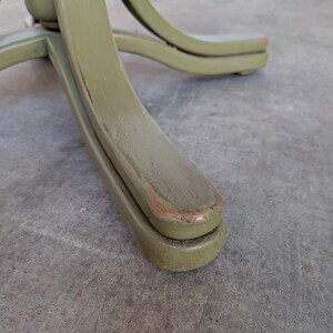 May include: Close-up of the base of a wooden chair. The chair base is painted a muted green color with visible wear and tear. The curved legs of the chair are visible, showcasing the design and construction of the furniture.