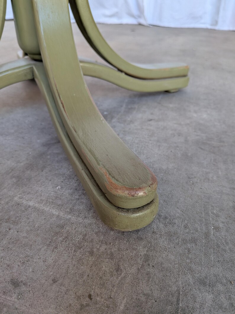 May include: Close-up of the base of a wooden chair. The chair base is painted a muted green color with visible wear and tear. The curved legs of the chair are visible, showcasing the design and construction of the furniture.