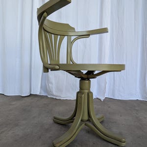 May include: An olive green vintage office chair with a curved back and armrests. The chair has a swivel base with a star-shaped footrest. The chair is made of wood and has a classic design.