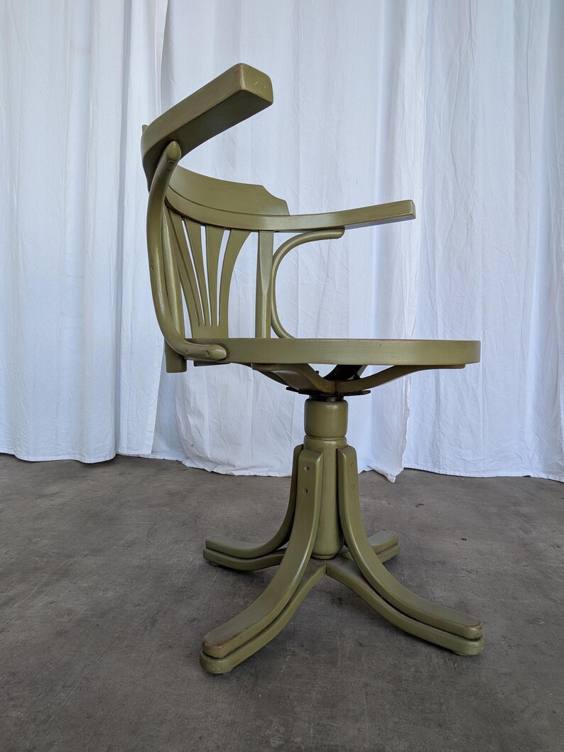 May include: An olive green vintage office chair with a curved back and armrests. The chair has a swivel base with a star-shaped footrest. The chair is made of wood and has a classic design.