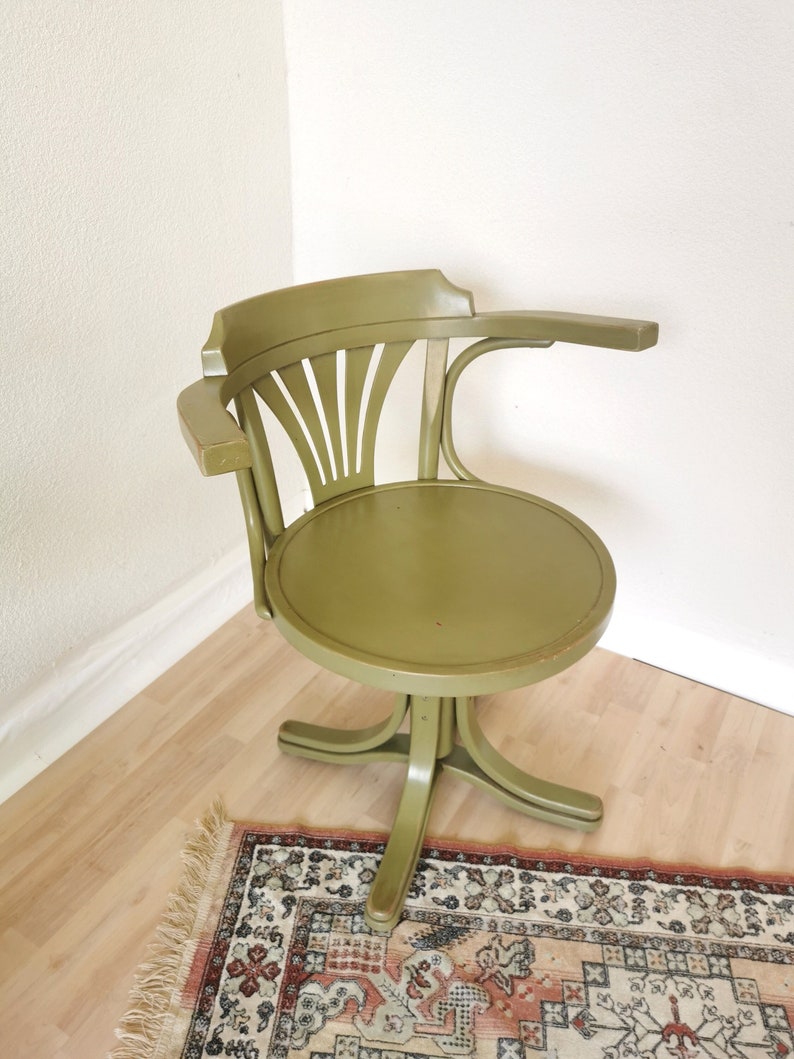 May include: An olive green vintage swivel chair with a curved back and armrest. The chair has a round seat and a five-point base. The chair is made of wood and has a classic design. The chair is placed on a light wood floor.