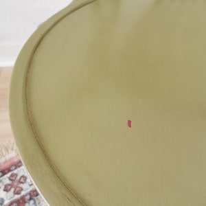 May include: Close-up of an olive green chair seat with a rounded edge. The chair has a curved back and legs, and a small red mark is visible on the seat. The chair appears to be made of wood and has a distressed finish.