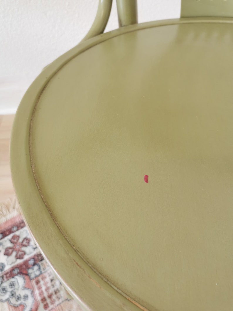 May include: Close-up of an olive green chair seat with a rounded edge. The chair has a curved back and legs, and a small red mark is visible on the seat. The chair appears to be made of wood and has a distressed finish.
