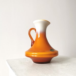 Könnte beinhalten: Eine vintage orangefarbene und weiße Glasvase mit einem ausgestellten Boden und einem Henkel. Die Vase hat eine glänzende Oberfläche und einen dezenten Ombre-Effekt.