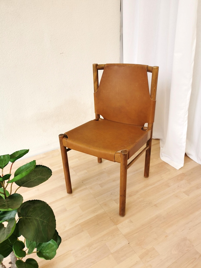 May include: A brown leather chair with a wooden frame. The chair has a simple design with a slightly curved back and a wide seat. The chair is sitting on a light wood floor.