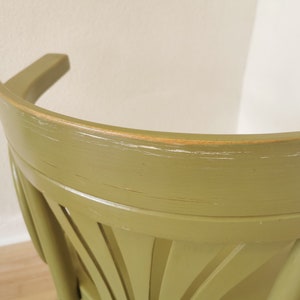 May include: Close-up of a green wooden chair back. The chair has a curved backrest and a decorative fan-like design on the lower back. The paint is slightly distressed, showing hints of the wood underneath.