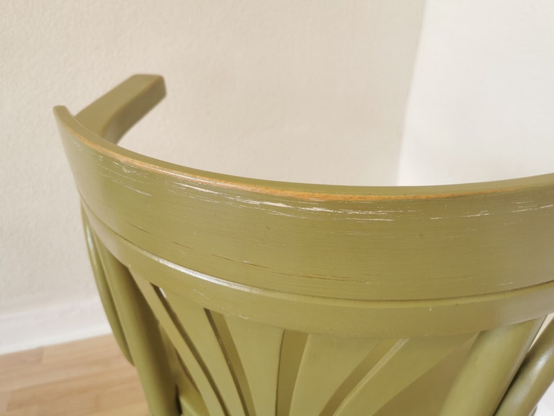 May include: Close-up of a green wooden chair back. The chair has a curved backrest and a decorative fan-like design on the lower back. The paint is slightly distressed, showing hints of the wood underneath.
