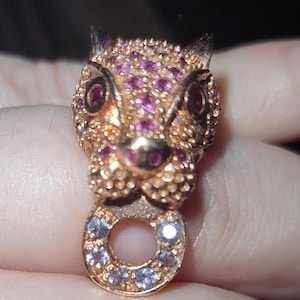 May include: A gold-coloured pendant shaped like a stylised cat's head, set with small, round, pink and clear gemstones. The pendant has a circular loop at the bottom, also decorated with gemstones. The cat's eyes are a deep red.