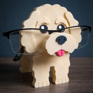 May include: A cream-colored 3D-printed dog figurine serving as an eyeglass holder. The dog has black eyes, a black nose, and a pink tongue. A pair of black-framed glasses rests on the dog's face. The text TIMUX is visible in the bottom right corner.
