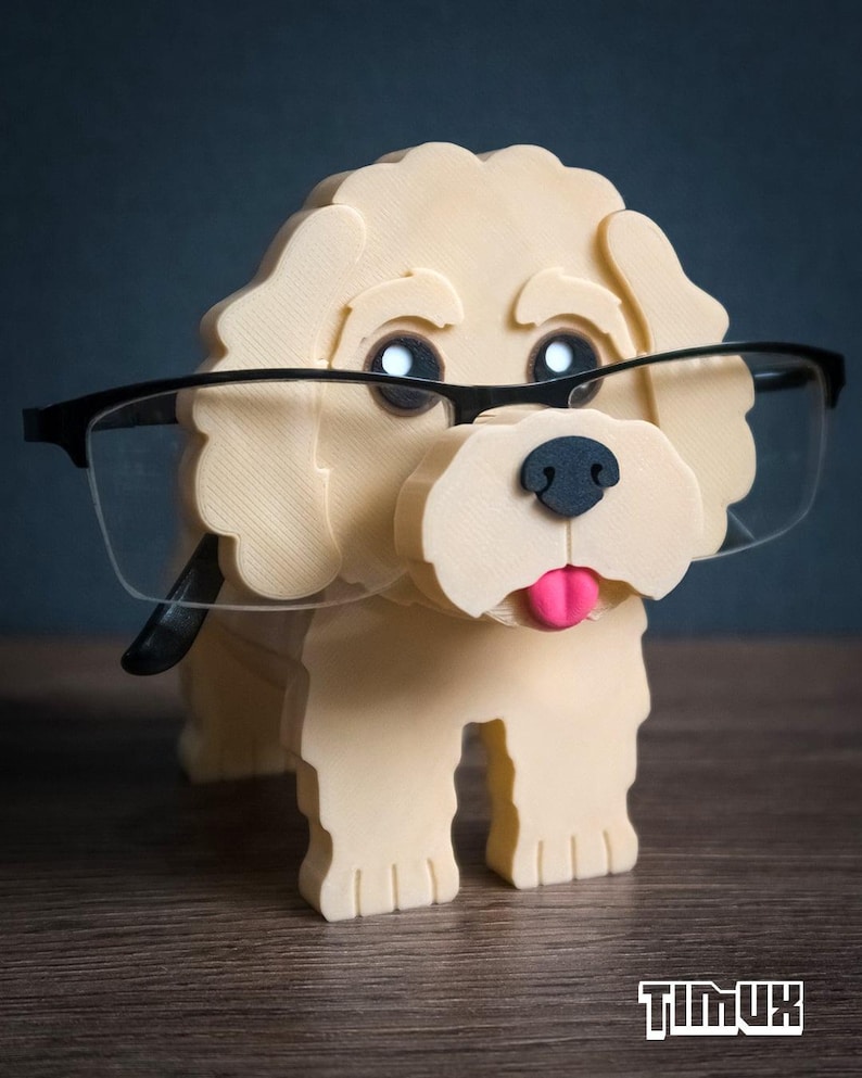 May include: A cream-colored 3D-printed dog figurine serving as an eyeglass holder. The dog has black eyes, a black nose, and a pink tongue. A pair of black-framed glasses rests on the dog's face. The text TIMUX is visible in the bottom right corner.