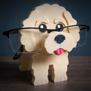 May include: A cream-colored 3D printed dog figurine serving as an eyeglass holder. The dog has a black nose, a pink tongue, and large dark eyes. A pair of black-rimmed glasses rests across the dog's face. The word TIMUX is visible at the bottom.