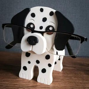May include: A novelty glasses holder in the shape of a Dalmatian dog. The dog is white with black spots and black ears. It is holding a pair of black-framed glasses. The holder is on a wooden surface.