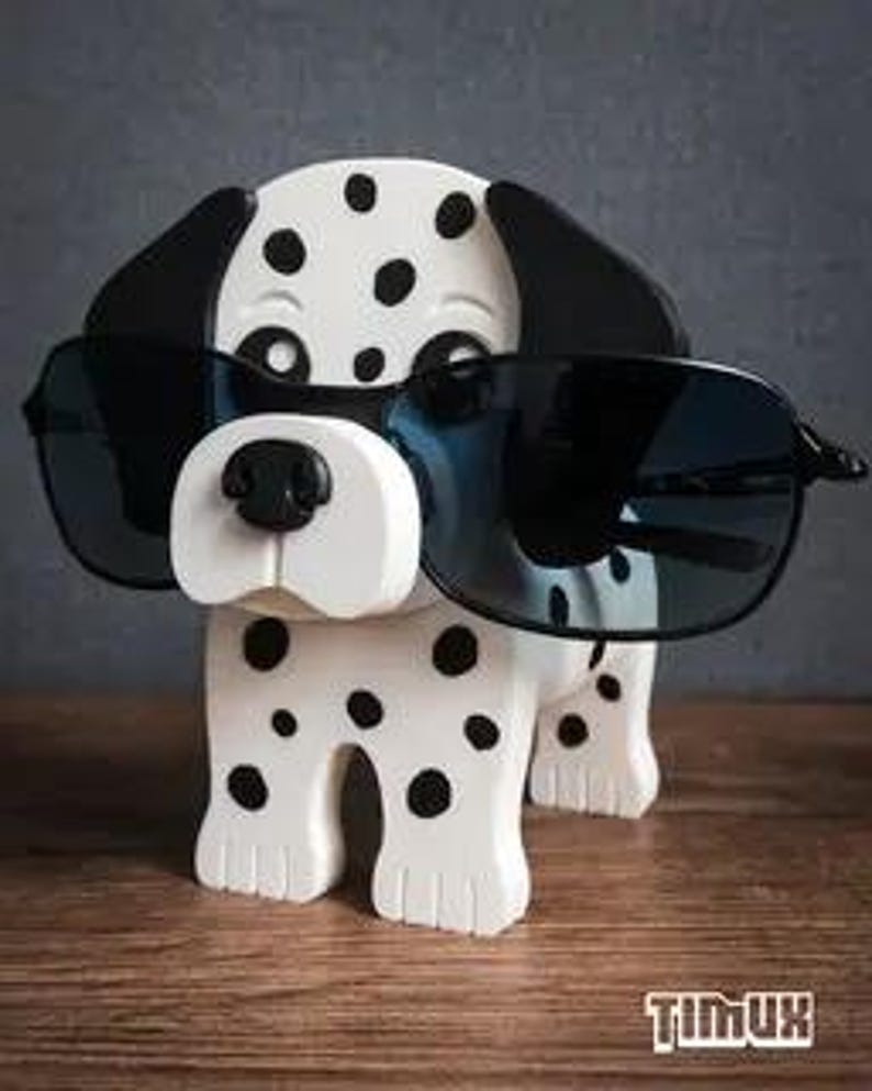 May include: A wooden Dalmatian dog ornament, white with black spots, wearing black sunglasses. The dog has a black nose and ears. The word "TIMUX" is printed on the bottom right of the ornament.