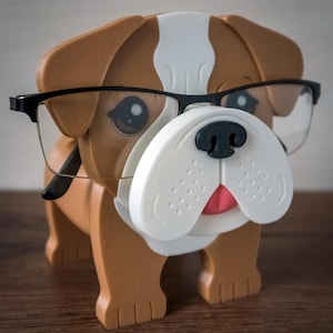 May include: A brown and white bulldog figurine wearing black framed glasses. The bulldog has a pink tongue sticking out and a black nose. The figurine is standing on a wooden surface.