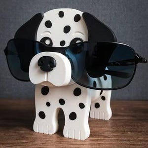 May include: A white and black spotted Dalmatian dog ornament wearing black sunglasses. The dog has black ears, nose, and spots. The sunglasses are black with dark lenses. The ornament is on a wooden surface.