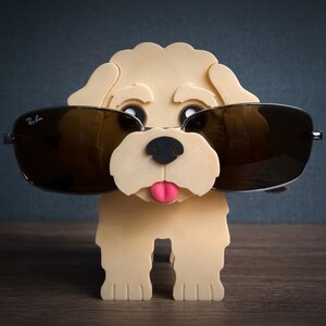 May include: A light beige 3D-printed dog figurine serving as an eyeglass holder. The dog has a black nose, a pink tongue, and is wearing dark-tinted Ray-Ban sunglasses. The figurine features a textured surface and stands on four legs.
