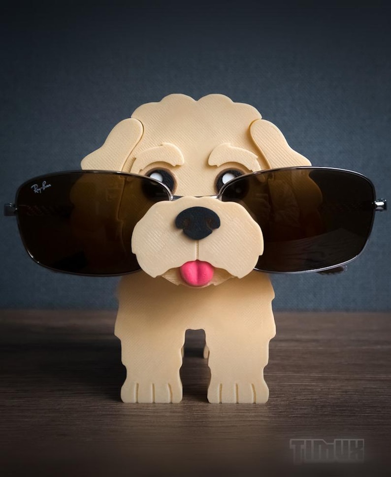 May include: A light beige 3D-printed dog figurine serving as an eyeglass holder. The dog has a black nose, a pink tongue, and is wearing dark-tinted Ray-Ban sunglasses. The figurine features a textured surface and stands on four legs.