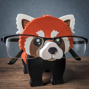May include: A whimsical red panda glasses holder with black-framed glasses. The holder is primarily orange, white, brown, and black, with a cute design. The glasses are positioned as if the panda is wearing them. The base has the word "TIMUX".