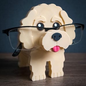 May include: A cream-colored dog-shaped eyeglass holder with a black nose, dark eyes, and a pink tongue. The figure features textured, layered construction and is wearing a pair of black-rimmed glasses. The word TIMUX is printed in white at the bottom right.
