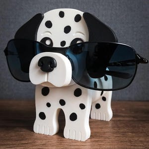 May include: A white and black spotted Dalmatian dog-shaped eyeglass holder wearing black sunglasses. The dog has black ears and a black nose. The eyeglass holder is on a wooden surface.