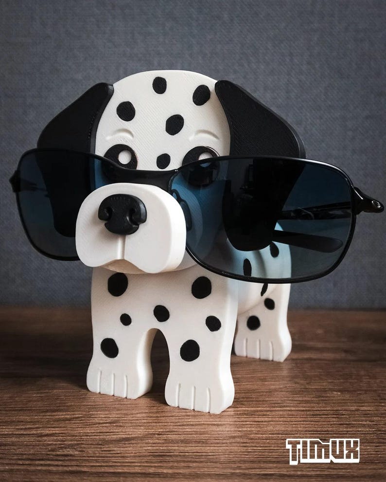 May include: A white and black spotted Dalmatian dog-shaped eyeglass holder wearing black sunglasses. The dog has black ears and a black nose. The eyeglass holder is on a wooden surface.