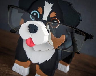 May include: A dog-shaped spectacle holder in black, white, and brown, wearing black-framed glasses. The dog has a red tongue and stands on a wooden surface. The brand name "TIMUX" is visible in the top right corner.