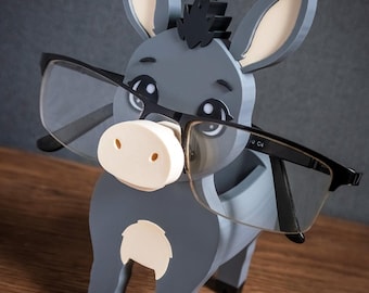May include: A gray donkey-shaped eyeglass holder with a cream-colored muzzle and inner ears, black hooves, and a black mane. The donkey is wearing black-rimmed glasses across its face. The text TIMUX is printed in white in the bottom corner.