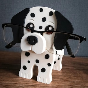 May include: A wooden eyeglass holder shaped like a Dalmatian dog, with black spots and ears. The dog is wearing black-framed glasses. The holder is white with black spots and sits on a wooden surface. The holder is a decorative item.