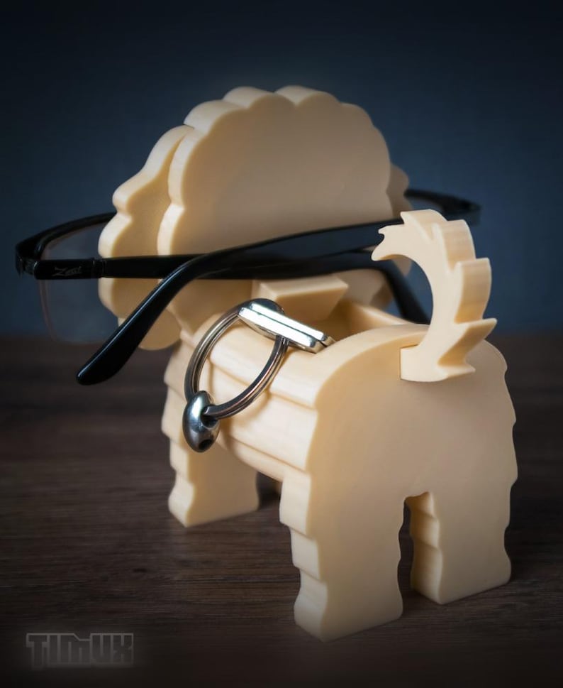 May include: A cream-colored, dog-shaped organizer made of layered material. The figure features a raised tail and a flat back section designed to hold a pair of black-framed eyeglasses. A silver-toned metal keyring rests on the dog's back.