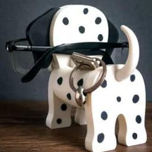 May include: A wooden Dalmatian dog-shaped eyeglasses and key holder. The dog is white with black spots and ears, holding a pair of eyeglasses. A key ring with a key is attached to the dog's nose. The brand name "TIMUX" is at the bottom.