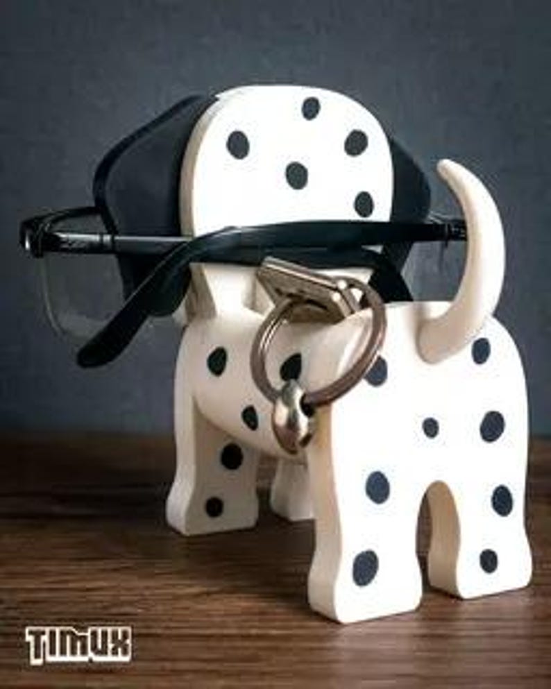 May include: A wooden Dalmatian dog-shaped eyeglasses and key holder. The dog is white with black spots and ears, holding a pair of eyeglasses. A key ring with a key is attached to the dog's nose. The brand name "TIMUX" is at the bottom.