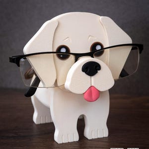 May include: A cream-colored dog-shaped eyeglass holder with a black nose, pink tongue, and brown eyes. The figure features a textured surface and is wearing a pair of black-rimmed glasses. The text TIMUX is printed in white in the bottom right corner.