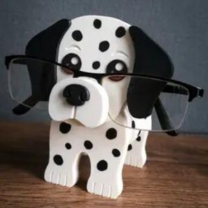 May include: A wooden eyeglass holder in the shape of a Dalmatian dog, with black spots and ears. The dog is holding a pair of black-framed spectacles. The holder has a white body and a black nose. The word "TIMUX" is printed on the bottom.