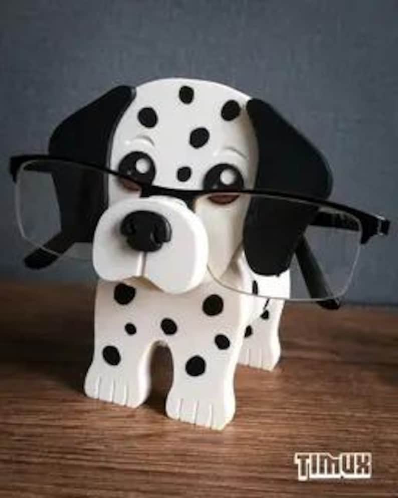 May include: A wooden eyeglass holder in the shape of a Dalmatian dog, with black spots and ears. The dog is holding a pair of black-framed spectacles. The holder has a white body and a black nose. The word "TIMUX" is printed on the bottom.