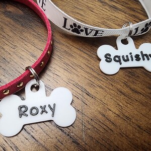 May include: Two pet collars with bone-shaped white acrylic tags. One collar is red suede with gold studs and a tag reading Roxy. The other is a white ribbon printed with the word Love and paw prints, featuring a tag reading Squishy.