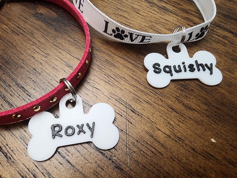 May include: Two pet collars with bone-shaped white acrylic tags. One collar is red suede with gold studs and a tag reading Roxy. The other is a white ribbon printed with the word Love and paw prints, featuring a tag reading Squishy.