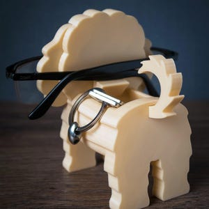 May include: A cream-colored, dog-shaped organizer made of layered material, featuring a slot for holding eyeglasses across the head and a small compartment for keys or accessories. The word TIMUX is printed in white at the bottom left corner.