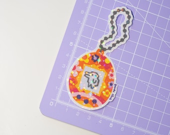 May include: A colorful sticker in the shape of a retro handheld game. The sticker features a pixelated character inside a square, surrounded by flowers and stars. The sticker has a holographic finish and a beaded chain.