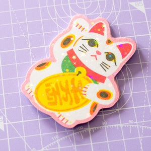 May include: A die-cut sticker of a Japanese Maneki-neko cat, a symbol of good luck. The cat is white with orange and black markings, a red collar, and a gold coin. The sticker has a pink border and holographic accents.
