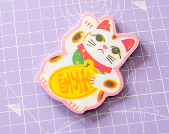 May include: A die-cut sticker of a white Maneki-neko cat with pink edges, featuring a red collar, green accents, and a yellow oval coin with red characters. The sticker has a holographic finish that reflects light in a star-like pattern.