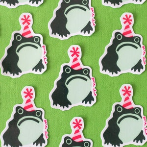 May include: A repeating pattern of frog stickers on a green backdrop. Each sticker depicts a dark green frog with a pale green face, wearing a red and white party hat. The text "garli club" is printed on each sticker.