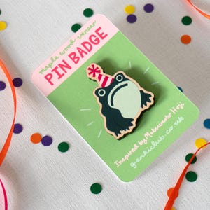 Könnte beinhalten: Ein Ansteck-Pin aus Ahornfurnier mit einem Cartoon-Frosch, der eine Partyhut trägt. Der Pin befindet sich auf einer grünen Karte mit dem Text "PIN BADGE". Bunte Konfetti und Bänder sind um den Pin herum verstreut.