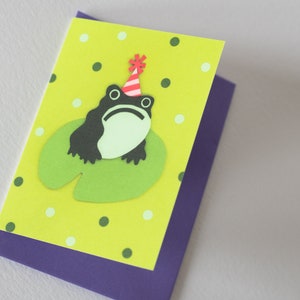May include: A handmade greeting card featuring a whimsical frog wearing a party hat, sitting on a lily pad. The card has a lime green background with white and dark green polka dots, and a purple backing.