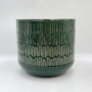 May include: A dark green ceramic plant pot with a textured leaf pattern. The pot has a rounded base and a cylindrical shape. The colour is a deep, forest green with lighter green accents in the textured design. Suitable for indoor plants.