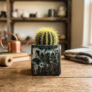 May include: A round, spiky green cactus with yellow needles planted in a square, textured ceramic pot. The pot features a mottled, dark green and black glaze with a rough, porous surface finish, sitting upright on a wooden surface.