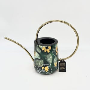 May include: A black watering can with a gold handle and a green and gold floral pattern. The can has a spout and a small tag attached to the handle.